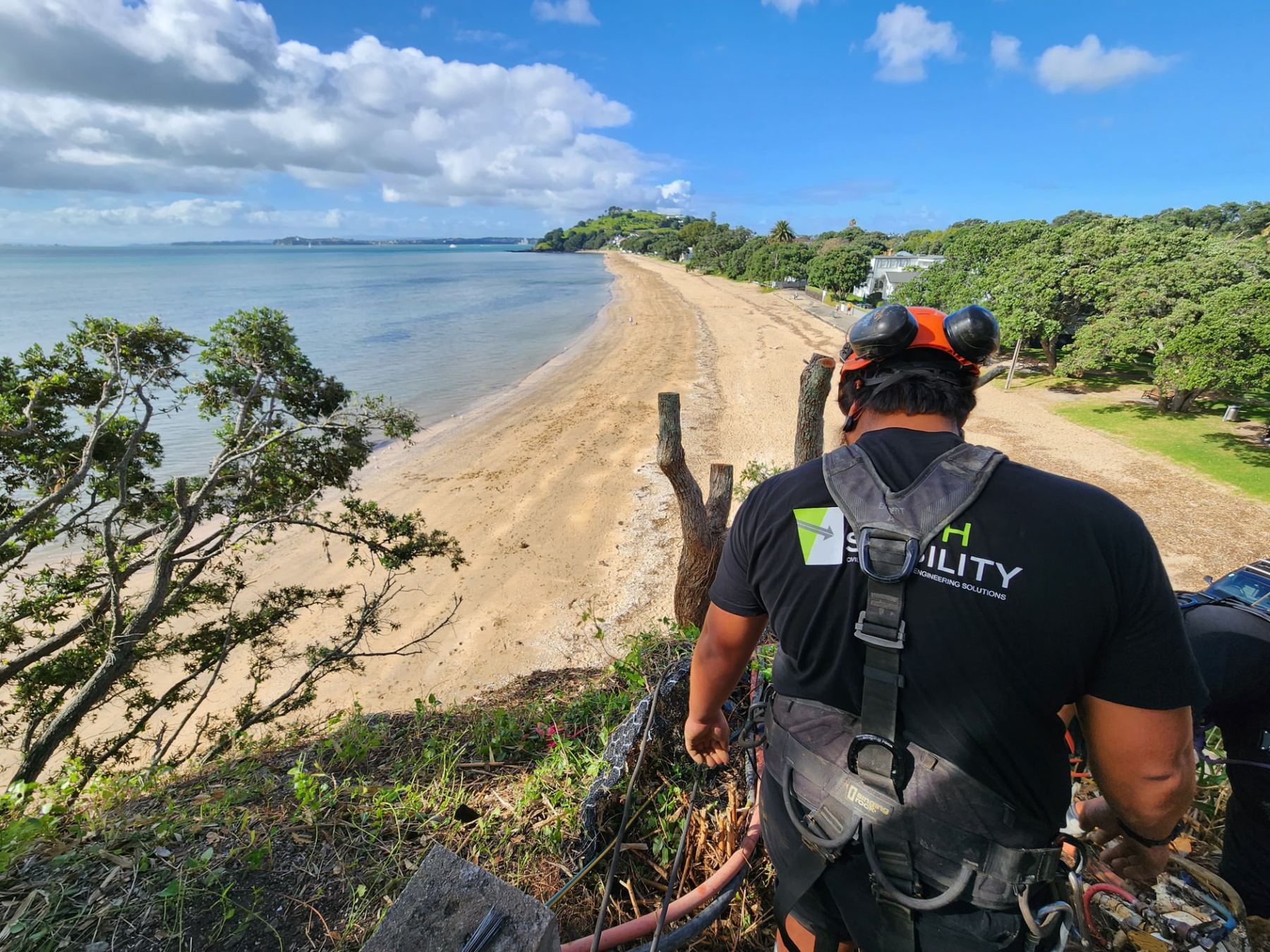 Technician in a safety harness standing at a grassy cliff edge overlooking Cheltenham Beach