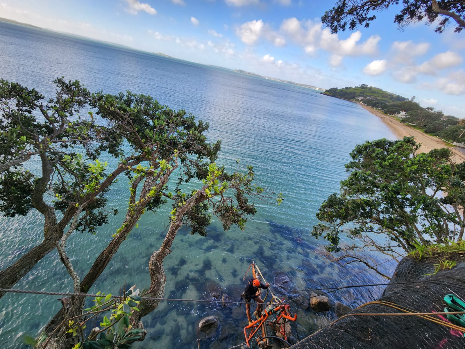 Rope access technician operating a drill rig on a cliff above the coast