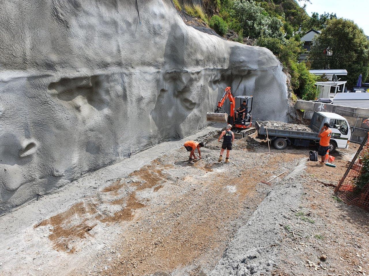 Excavator shaping a freshly sprayed shotcrete slope behind a property