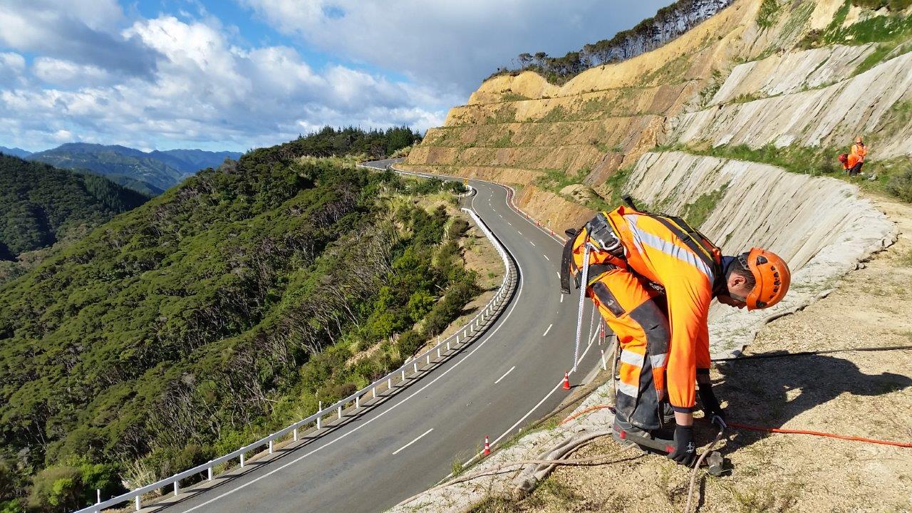 Workers in safety gear tending a stabilised slope above a winding mountain road