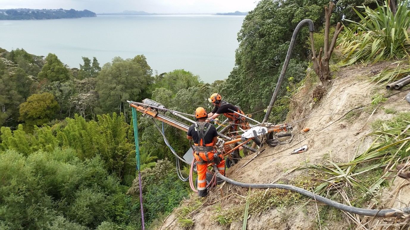 Technicians operating a drill rig on a steep cliff top above the harbour
