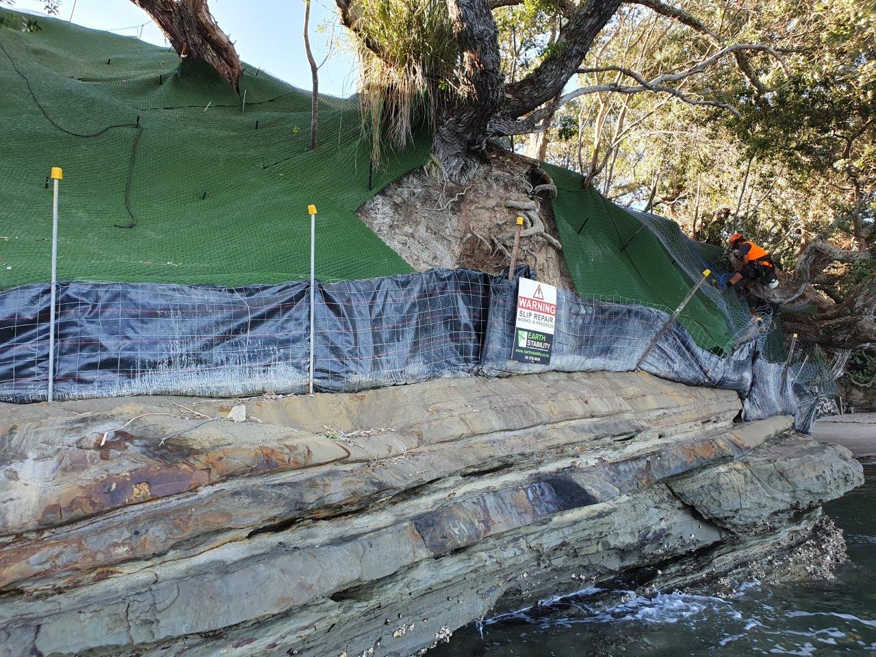 Silt fence and warning signage on a coastal rock platform below erosion matting