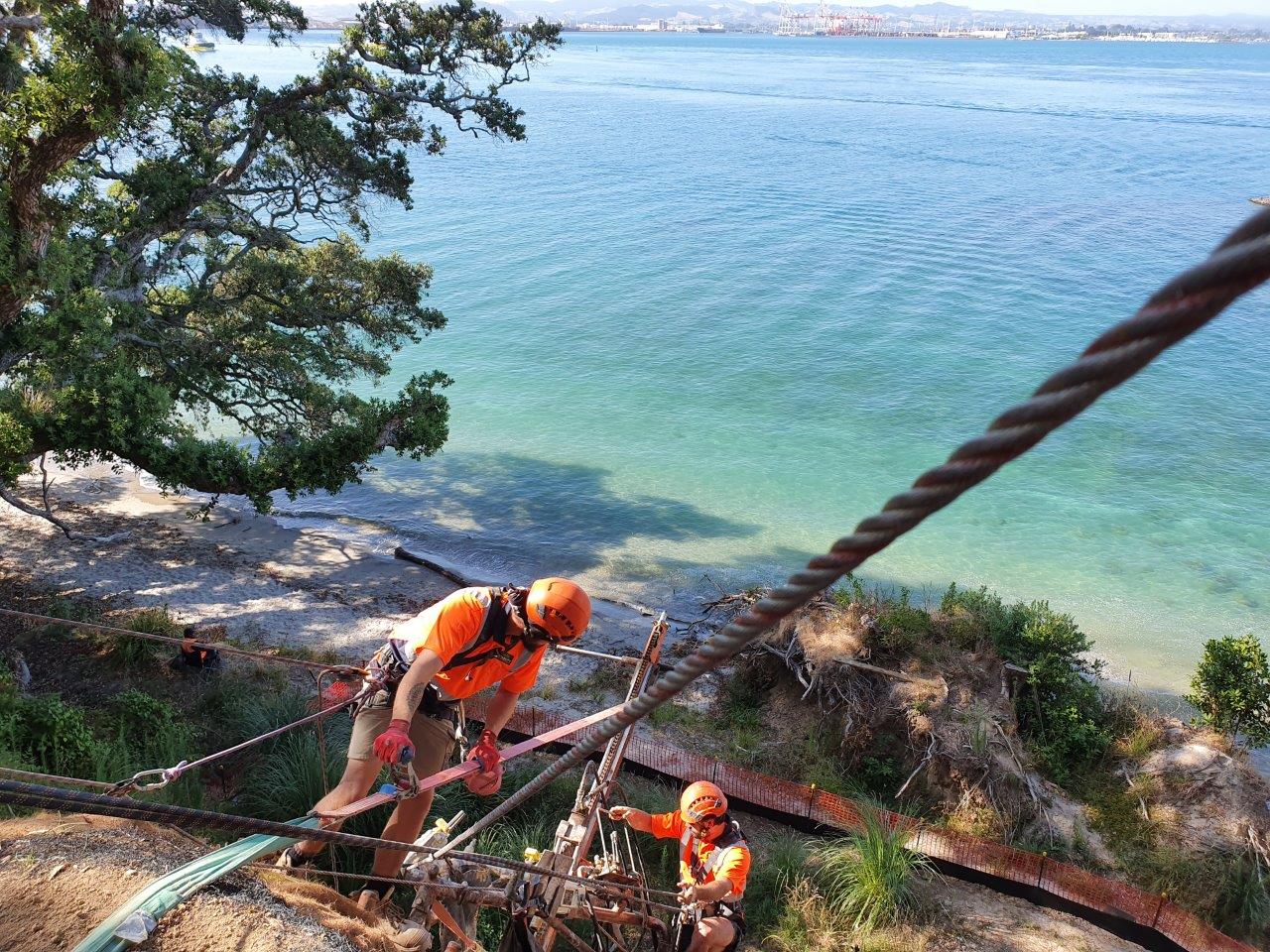 Two rope access technicians drilling on a cliff above turquoise water