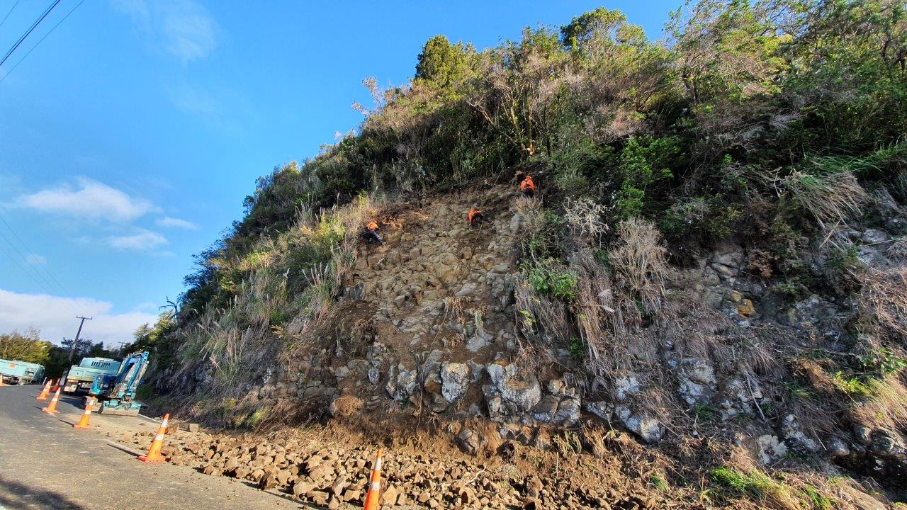 Rope access crew scaling loose rock from a roadside cut above traffic cones