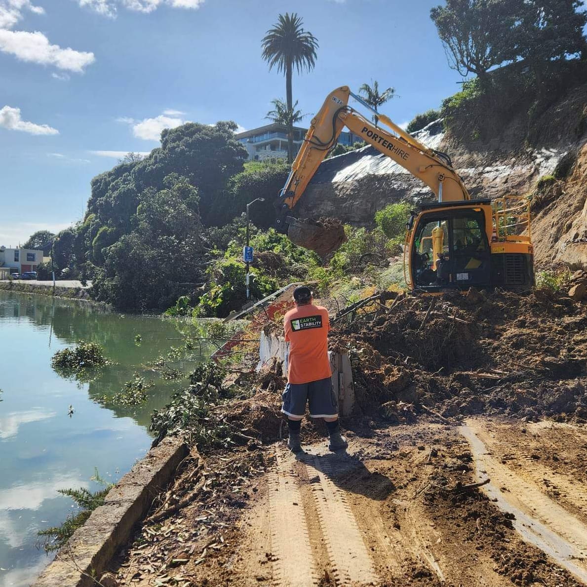Excavator clearing slip material on a coastal margin while a crew member looks on
